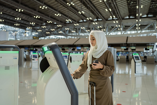 Happy muslim women at self check in desk in airport, holds passport, going to register for flight.