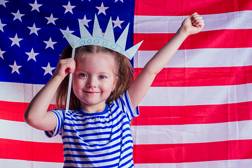 redhead blonde charming female kid celebration independence holding a paper torch and diy crown and american flag on a pink background in the studio.English language learning concept and freedom