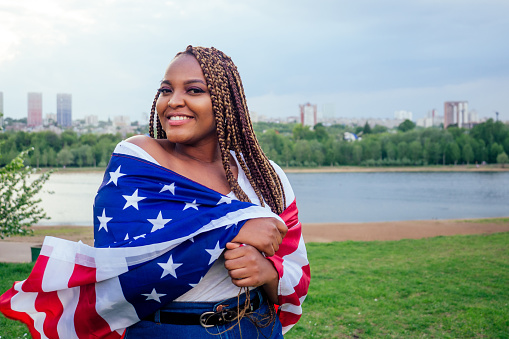 Smiling afro american woman holding USA flag and looking at camera autumn evening spring in the park by the lake