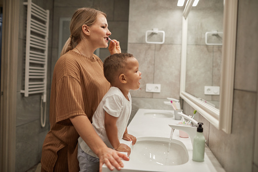 Side view portrait of young mother helping cute toddler boy brushing teeth in bathroom, copy space