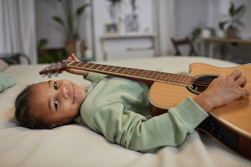 Portrait of cute black girl holding guitar and looking at camera while lying on bed at home, copy space