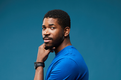 Portrait of handsome black man looking at camera over shoulder with smug expression while wearing blue on blue background, copy space