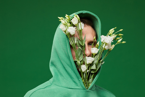 Portrait of young man with white Spring roses in hood of shirt against vibrant green background, copy space