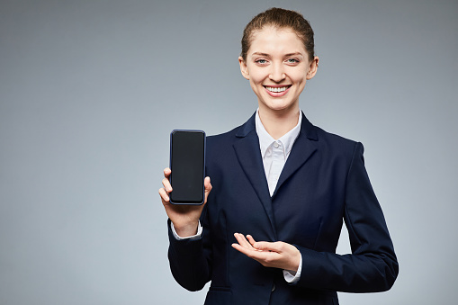 Waist up portrait of smiling businesswoman holding smartphone with blank screen to camera against grey background, copy space