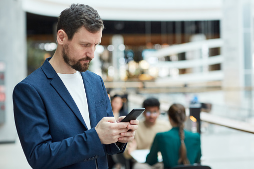Waist up portrait of adult bearded man using smartphone in shopping mall and wearing business jacket, copy space