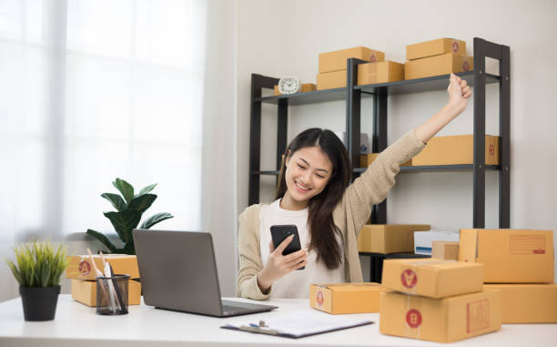 Happy Young asian entrepreneur woman checking product on smartphone. Business woman working SME online at home. Check address on parcel. Small business owner start up freelance concept. feliz joven empresaria asiática revisando el producto en el teléfono inteligente. mujer de negocios que trabaja pymes online en casa. verifique la dirección en el paquete. propietario de una pequeña empresa que inicia el concepto independiente.