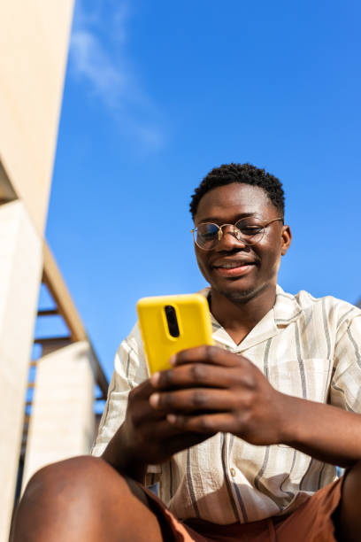 black young man sitting on stairs outdoors using mobile phone. vertical image. copy space. - homework teenager mobile phone school imagens e fotografias de stock