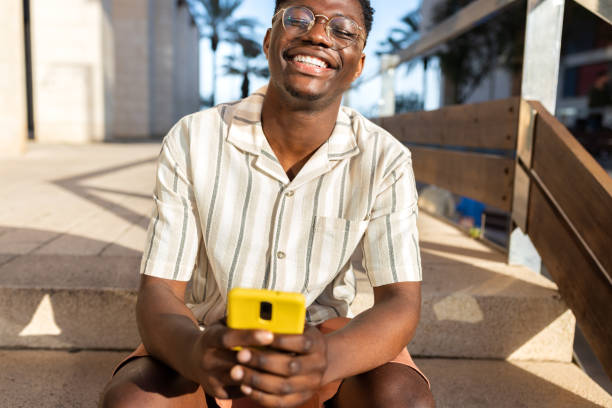 istock Happy black man sitting on stairs, relaxing outdoors using mobile phone, laughing. 1403955739