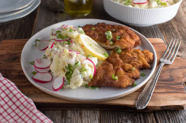 Traditional german, bavarian dish with homemade fresh made potato salad with red radish and pan fried and breaded schnitzel. Served on a plate with lemon on rustic and wooden table background.