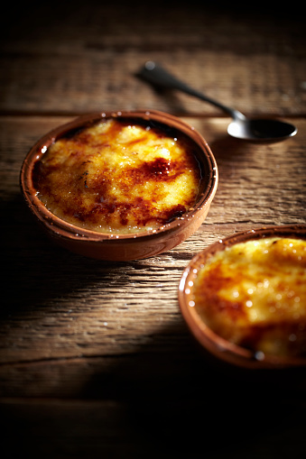 Two small bowls of crème brûlée on a wooden table. The bowls are brown and there is a spoon on the table.