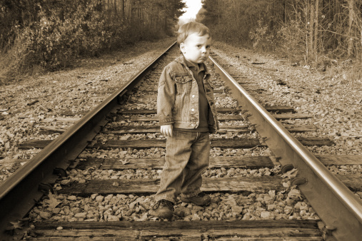 Serious looking two year old boy on rail road tracks in the country wearing blue jeans, blue jean jacket, and boots.  Image is in sepia.  