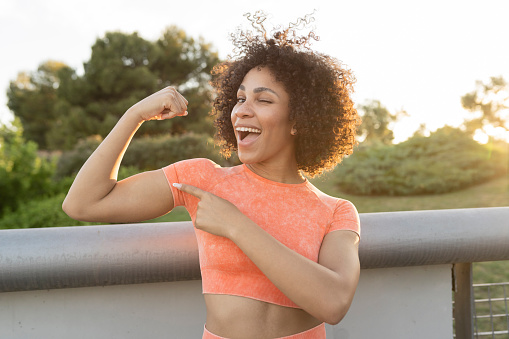 portrait of a beautiful African American woman wearing sportswear, showing her biceps and looking at the camera winking happily, outdoors at sunset