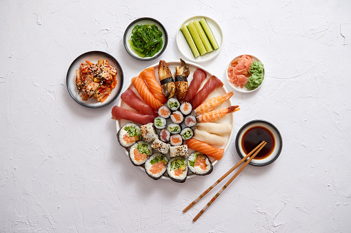 Asian food assortment. Various sushi rolls placed on round ceramic plate. Kimchi and goma wakame salads, fresh cucomber, ginger and wasabi. Soy souce and chopsticks on sides.