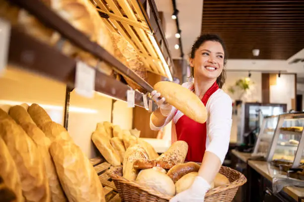 Photo of Bakery female worker in uniform selling loaf of bread to the customer in bakehouse. In background shelf full with fresh pastry products for sale.