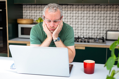 A worried man touches his face in an anxious way after looking at bad news on his laptop.