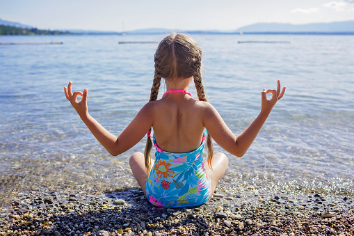 Cute girl has a rest and sits in a lotus pose on pebble beach at Geneva Lake over blue mountains in sun light, daydreaming and happiness, summer vacations, travel and summertime, lifestyle, back view
