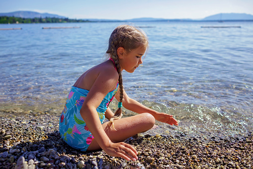 Happy girl has fun and plays with stones on pebble beach at Geneva Lake over blue mountains in sun light, lots of fun and happiness, summer vacations, travel and summertime, lifestyle