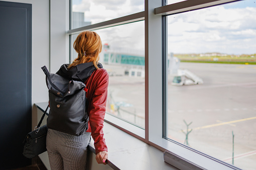 Rear view on woman waiting in departure area and looking at airport runway through the window