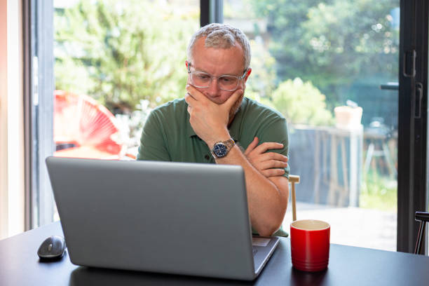 обеспокоенный мужчина смотрит на ноутбук в современном домашнем интерьере - uk check finance home finances стоковые фото и изображения
