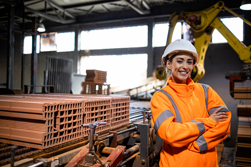 Portrait of female production line worker in safety equipment standing by conveyer machine transporting clay bricks in factory.