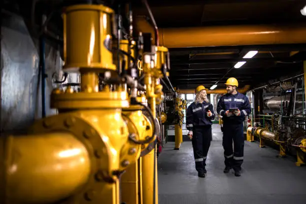 Photo of Oil and gas refinery production. Factory workers in safety equipment walking by gas pipes and checking distribution and consumption.