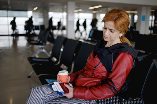 Female passenger waiting in airport departure area, holding passport in hand and coffee to go