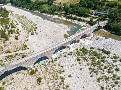 Aerial view. Drought and dry rivers. Roman bridge of Bobbio over the Trebbia river, Piacenza, Emilia-Romagna. Italy. 06-16-2022. River bed with stones and vegetation. Called hunchback bridge, old or devil's bridge. Bathers.