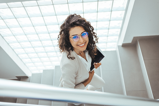 Shot of a young businesswoman using a digital tablet while walking up a staircase in an office. Businesswoman ascending office staircase. Professional Businesswoman Walking on Stairs in an Office Lobby