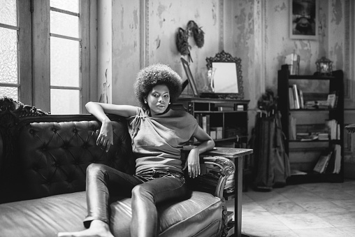 beautiful young black woman sitting on sofa in old house in havana, looking at camera