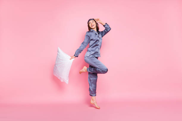 foto de tamaño corporal de cuerpo entero de una niña con ropa de noche que mantiene la almohada saltando mirando espacio en blanco aislado sobre fondo de color rosa pastel - ropa de dormir fotografías e imágenes de stock