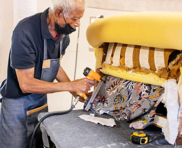 Senior male worker fixes the fabric with a pneumatic stapler on the body of sofa in the upholstery factory