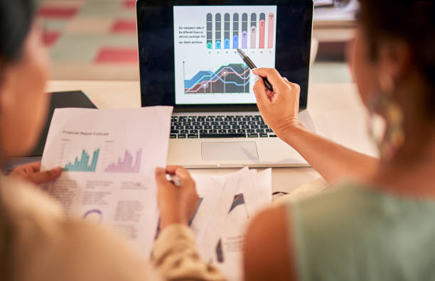 istock Two unrecognizable businesswomen sitting at their desks working on a laptop with documents. Female developers planning their business and discussing graphs and admin paperwork. Innovative entrepreneur 1403997263