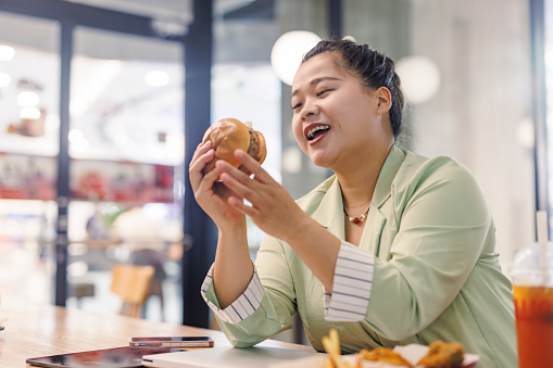 A plump Asian business woman happily eats hamburgers in a fast food restaurant.