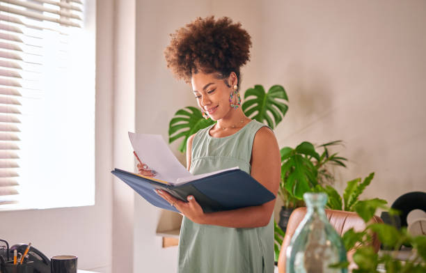 una giovane donna d'affari di razza mista che guarda un portafoglio in ufficio in piedi accanto a una finestra. una giovane sviluppatrice africana che legge documenti. azienda di design imprenditoriale innovativo - portfolio foto e immagini stock