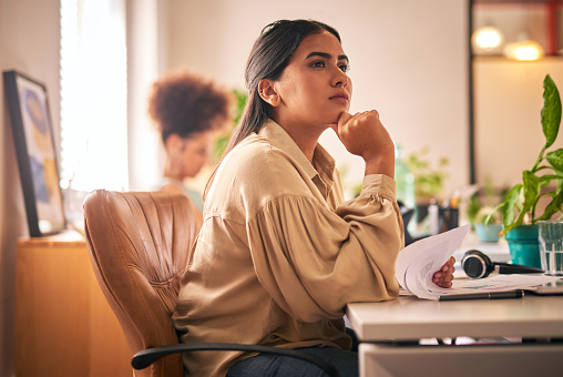 One young mixed race businesswoman working at her office desk. One young female indian developer looking away thoughtfully. Thinking about her ideas and planning for successa