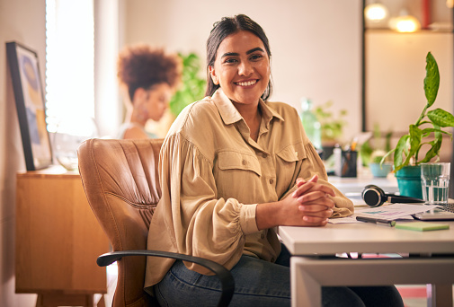 Portrait of mixed race businesswoman working at her laptop sitting at her desk in the office. One young female indian developer looking at the camera and smiling. Innovative entrepreneur design firm.
