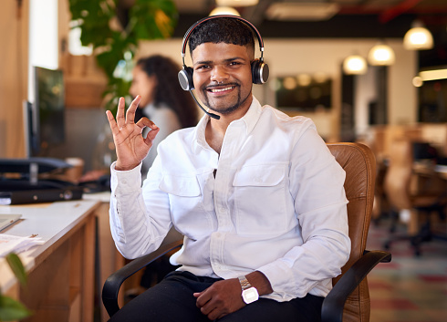 Call centre agent smiling with a-okay hand gesture. Portrait of a happy young man using a headset in a modern office.