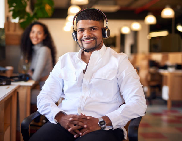 istock Call centre agent smiling while wearing a headset in a modern office. Professional, confident man sitting at his desk 1404005581