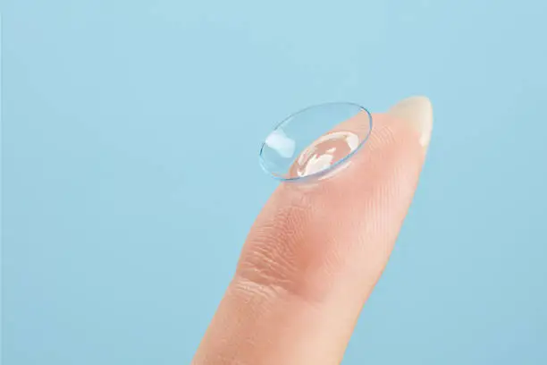 Contact lens on female finger on a blue background, close up. Medicine and vision concept. Woman holding soft contact lens. Shallow depth of field.