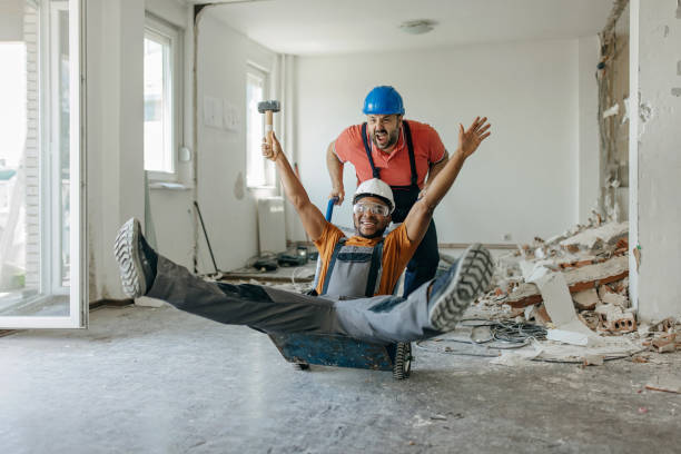 An African American male construction worker is smiling wide, while sitting on a construction trolley, pushed by his Caucasian coworker.