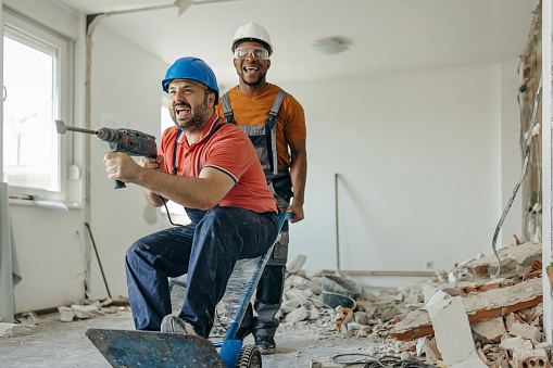 Two multiracial construction workers are being playful at work, while the Caucasian man is holding a pneumatic sledgehammer in his hands.