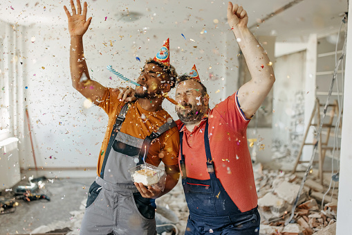 An African American and a Caucasian male construction worker are celebrating their new project with cake and confetti.