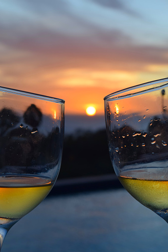 Two wine glasses, filled with white wine, toasting with each other as the sun sets over the pool and the Caribbean sea.