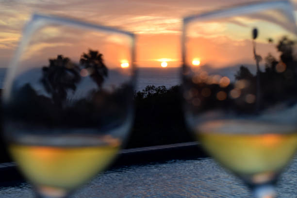Two wine glasses, filled with white wine, toasting with each other as the sun sets over the pool and the Caribbean sea.