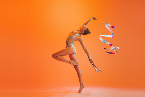 Dance, emotions. Young sportive girl, rhythmic gymnastics artist dancing isolated on orange color background. Concept of sport, action, aspiration, education, active lifestyle