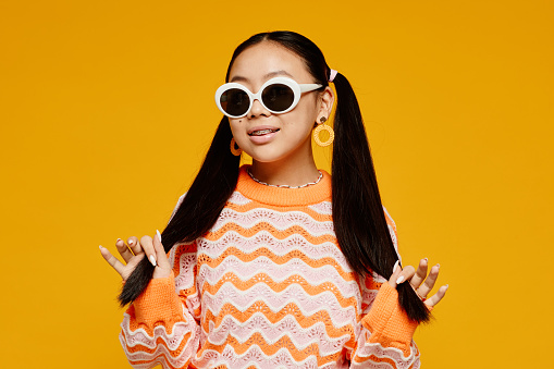 Waist up portrait of teenage Asian girl with pigtails wearing white sunglasses over vibrant yellow background
