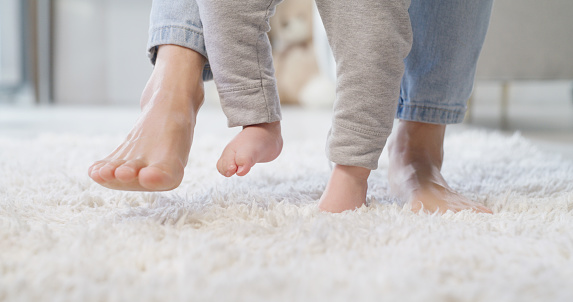 Closeup of parent and child playing and dancing on a carpet at home. Mother and baby feet walking in a bright white living room. Modern single parenting and child care