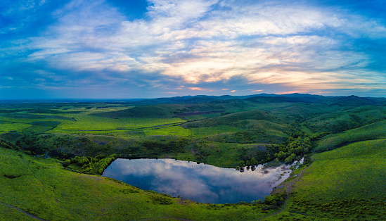 Panorama on a small swampy mountain lake with clear dark cool water is located between the hills greened with fresh spring new grass, in the Balkan mountains under a cloudy blue sunny sky