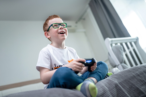 Cute joyful red-haired boy with a gamepad playing video games sitting on a bed in bedroom