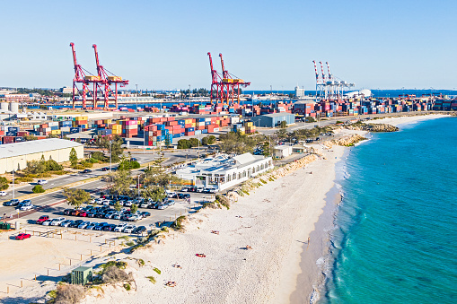 Nature & Industry: aerial sunny view of Fremantle's Port Beach with nearby busy container port; people sunbathing on white sandy beach; turquoise-coloured water, Coast entertainment venue in centre frame with crowded carpark; stacks of multi-coloured containers on hard stand in port facility with large gantry cranes standing idle. Juxtaposition, nearby, cooperation, close proximity